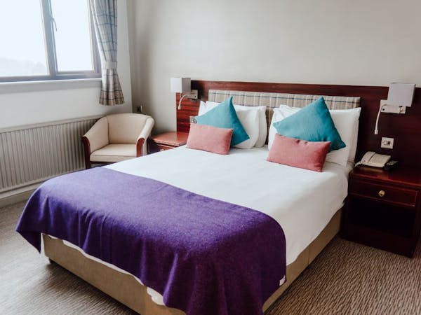 A cosy hotel bedroom with a comfortable bed with tweed cushions and throw, chair and a window with tweed curtains.