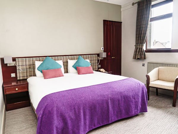 A cosy hotel bedroom with a comfortable bed with tweed cushions and throw, chair and a window with tweed curtains.