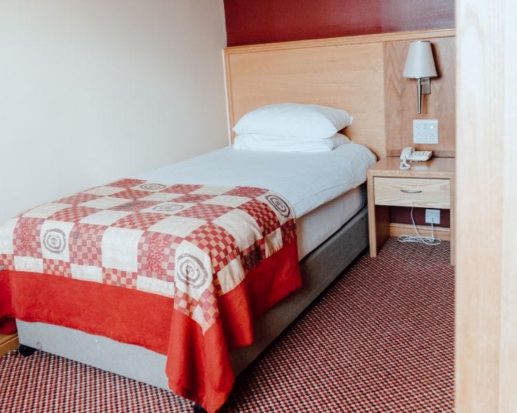Single room with white bed linen and red check bed runner, side table holds telephone and wall mounted lamp