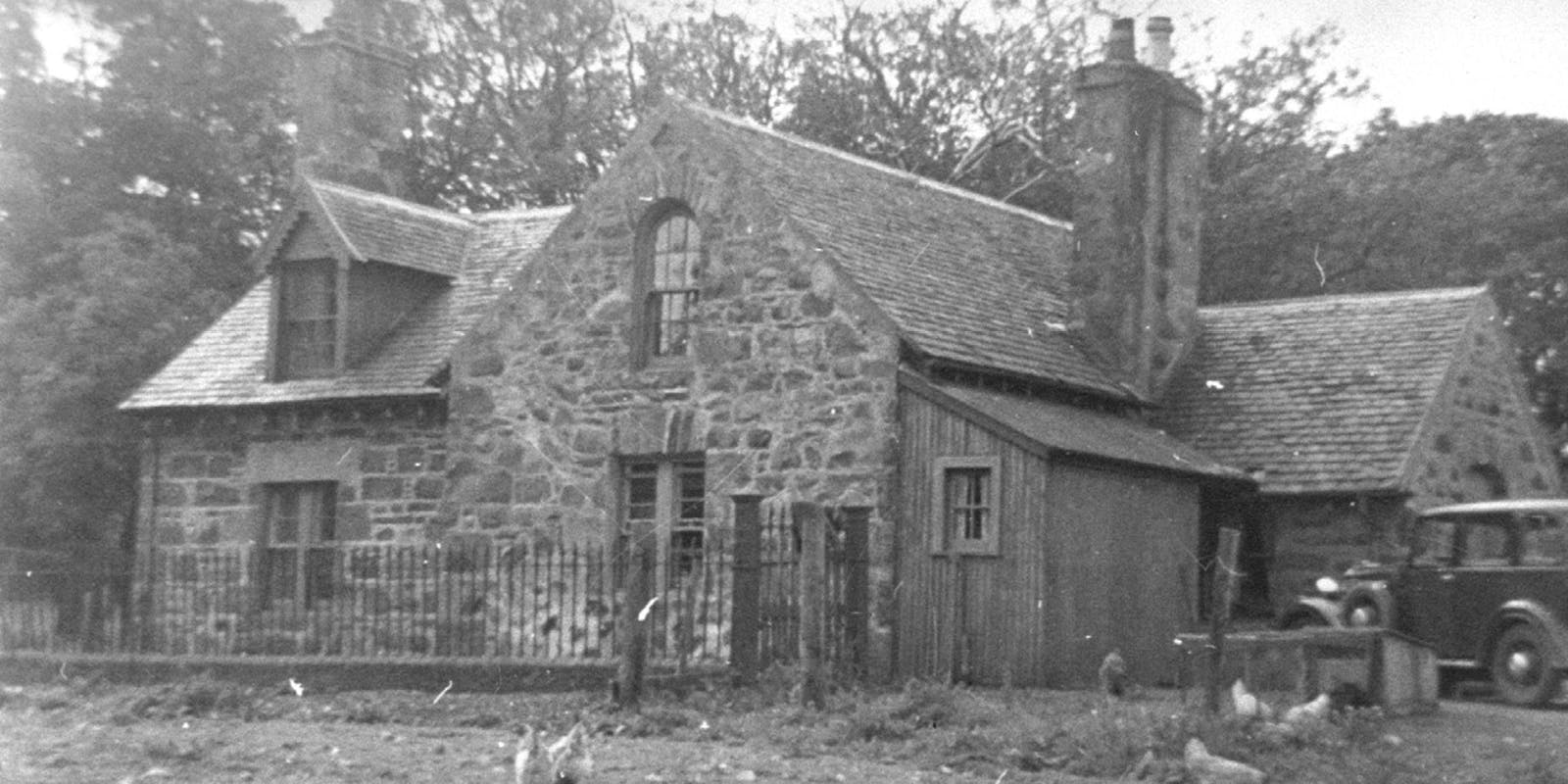Black-and-white photo of a manor farmhouse with chimneys, fenced yard, wooden extension, and an old car parked to the right.