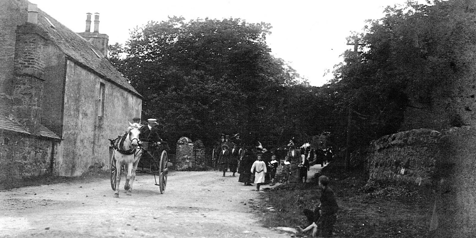 Black and white photo of a horse-drawn carriage on a dirt road passing a group of people near stone buildings and trees.