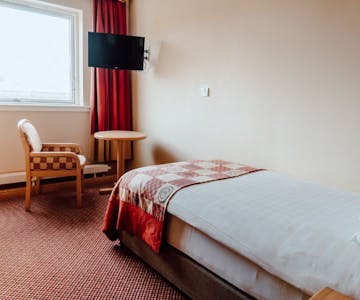 Single room with white bed linen and red check bed runner and complimenting chair. Wall mounted and window on outside wall.