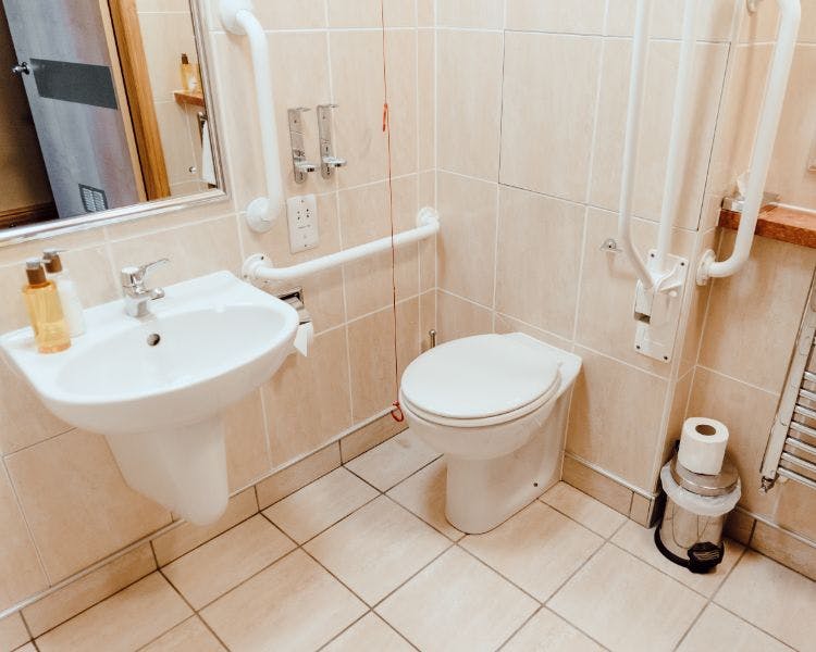 Accessible bathroom with neutral tiling, handrails. pull cord shower and white sink.