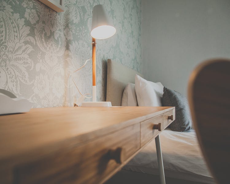 A desk with a switched on table lamp standing in front of a single bed