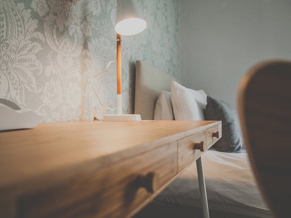 A desk with a switched on table lamp standing in front of a single bed