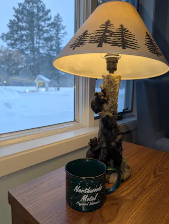 Image from inside looking out at a snowy backdrop. The image has a Northwoods lamp and a Northwoods Motel coffee mug