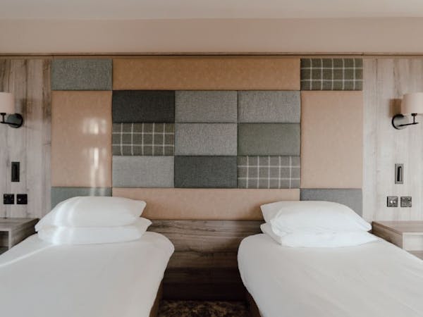 Twin beds in a hotel room with a tweed and leather fabric covered accent wall.