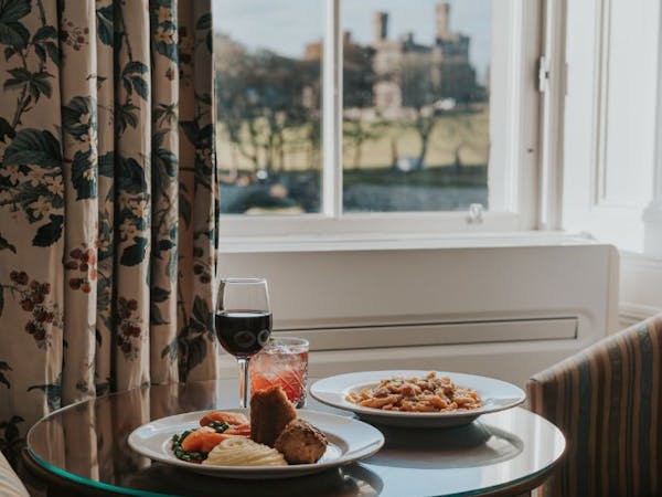 A table set with two plates of food and glasses of wine, placed in front of a window with views over the marina to castle.