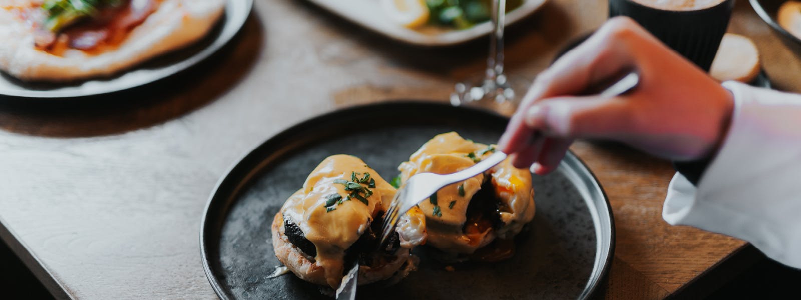 A person slicing into a poached egg with hollandaise sauce on a plate, preparing to enjoy the dish.