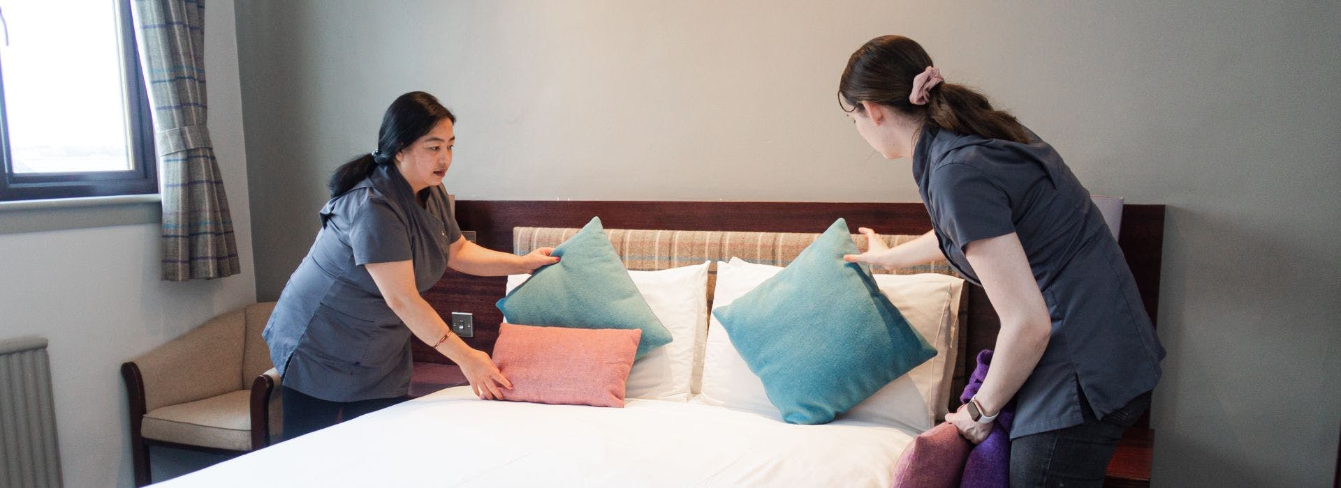 Two housekeepers making a bed with white linen and tweed cushions in blue and pink