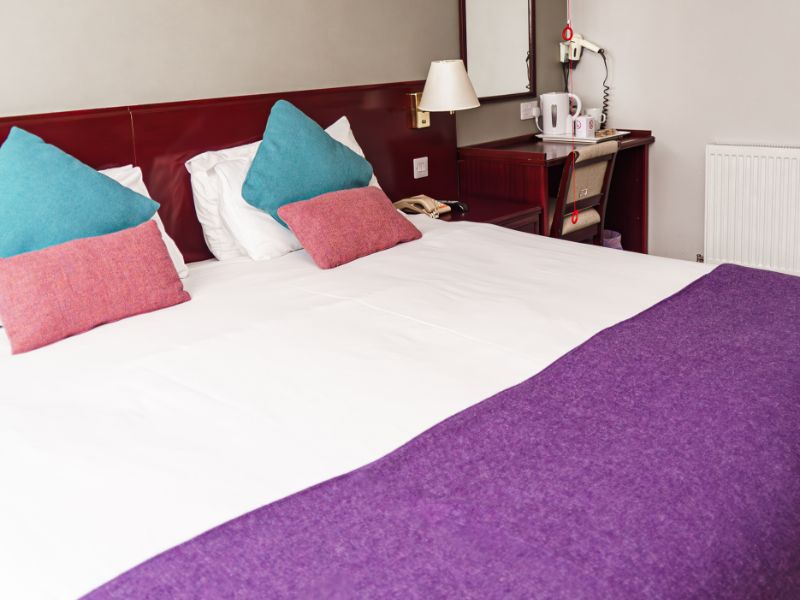 A cosy hotel bedroom with a comfortable bed with tweed cushions and throw, chair and a window with tweed curtains.