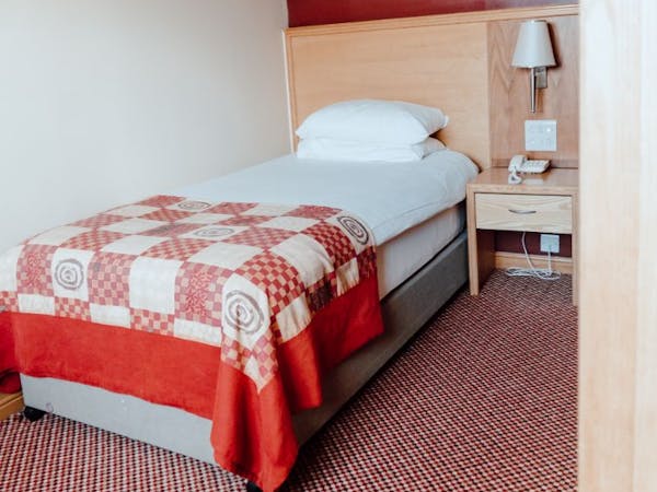Single room with white bed linen and red check bed runner, side table holds telephone and wall mounted lamp