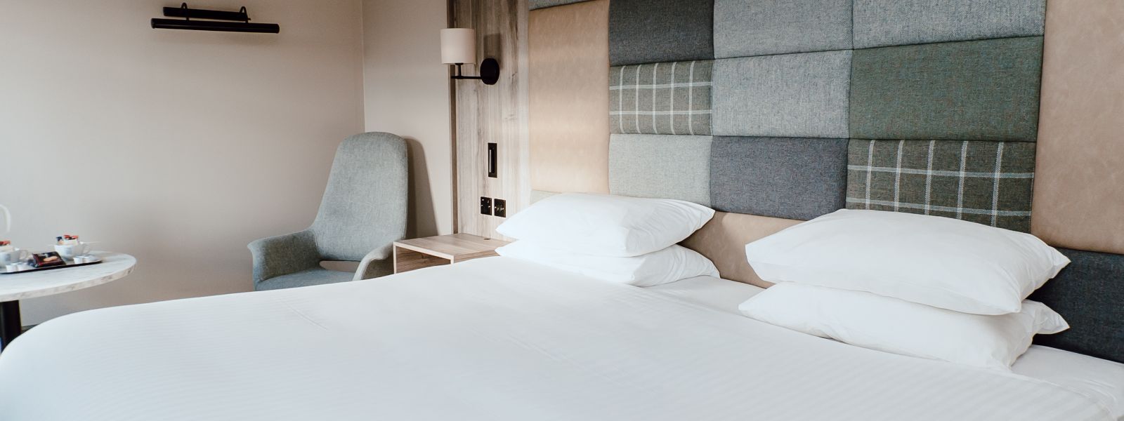 Double bedroom with white linen, check and plain tweed patchwork feature headboard, wall mounted lamps.