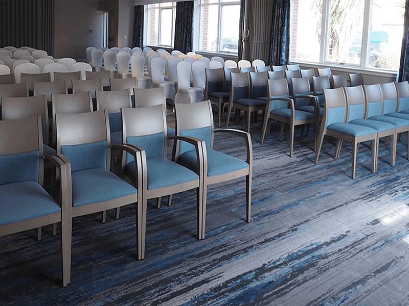 Theater style function room with blue, grey chairs and carpet, windows to the left with complimentary coloured curtains.