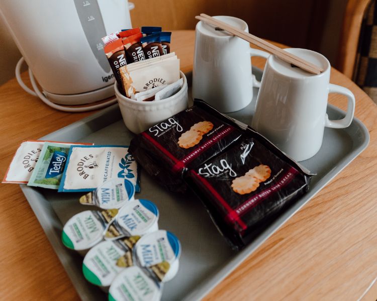 Tea and coffee station setup with kettle, cups, milk and biscuits on a tray placed on a wood table.