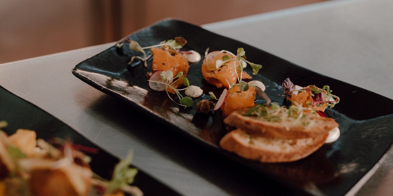 Smoked salmon and micro herb appetiser on a black plate sat on a dark table.