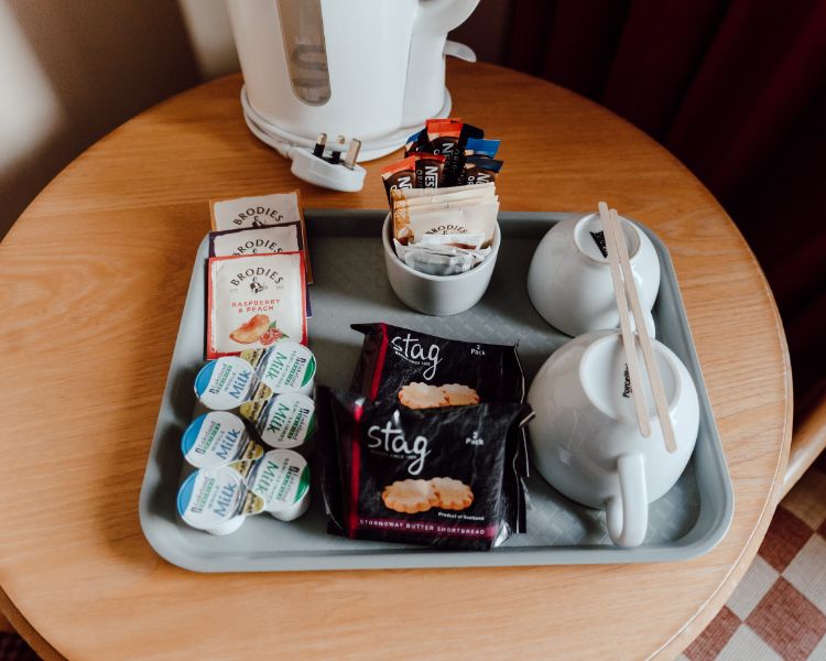 Tea and coffee station setup with kettle, cups, milk and biscuits on a tray placed on a wood table.