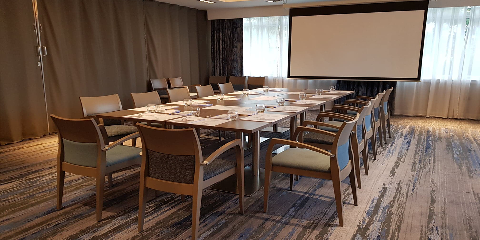 Table meeting space with OHP screen, blue, grey chairs and carpet, windows to the back with complimentary coloured curtains.