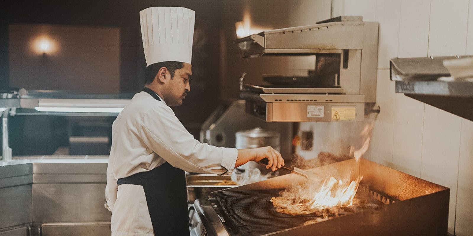 A chef wearing a white uniform and a tall white hat uses tongs to grill food over an open flame in a commercial kitchen.