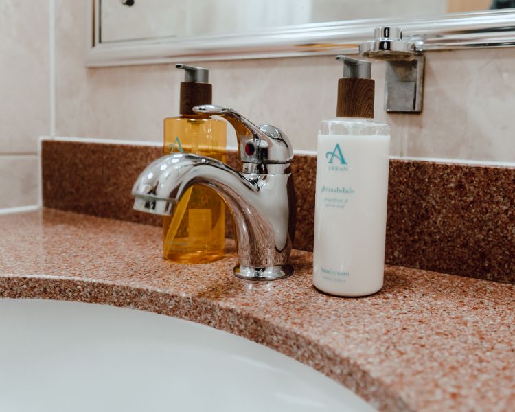 Bathroom closeup with fixed tap on brown sink suround and soap and hand cream dispensers.