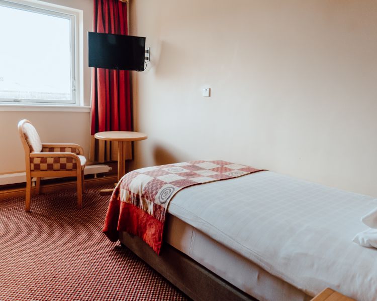 Single room with white bed linen and red check bed runner and complimenting chair. Wall mounted and window on outside wall.