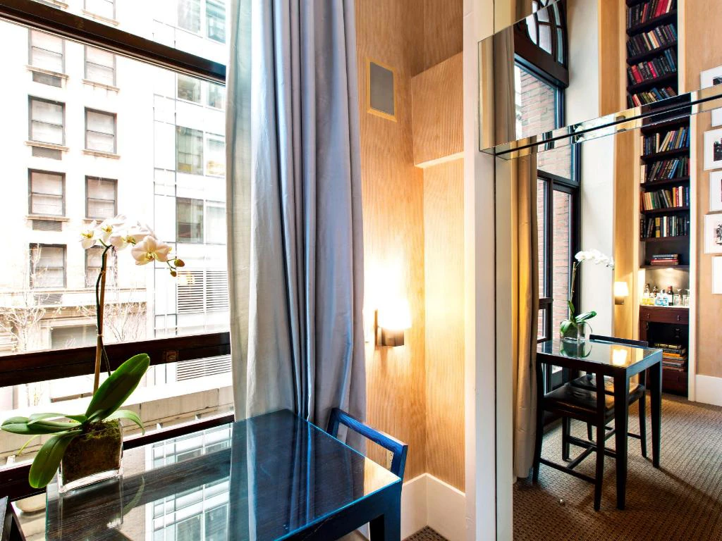 A hotel room with a glass desk, blue chair, and orchid plant near a window overlooking the city.