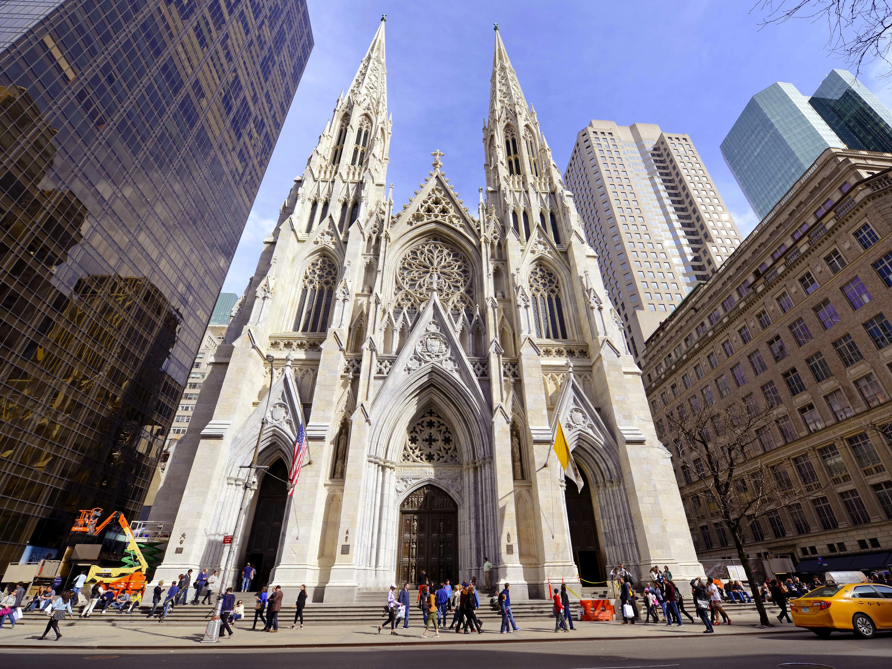 A large cathedral with two spires and a Gothic facade surrounded by skyscrapers and people walking on the sidewalk