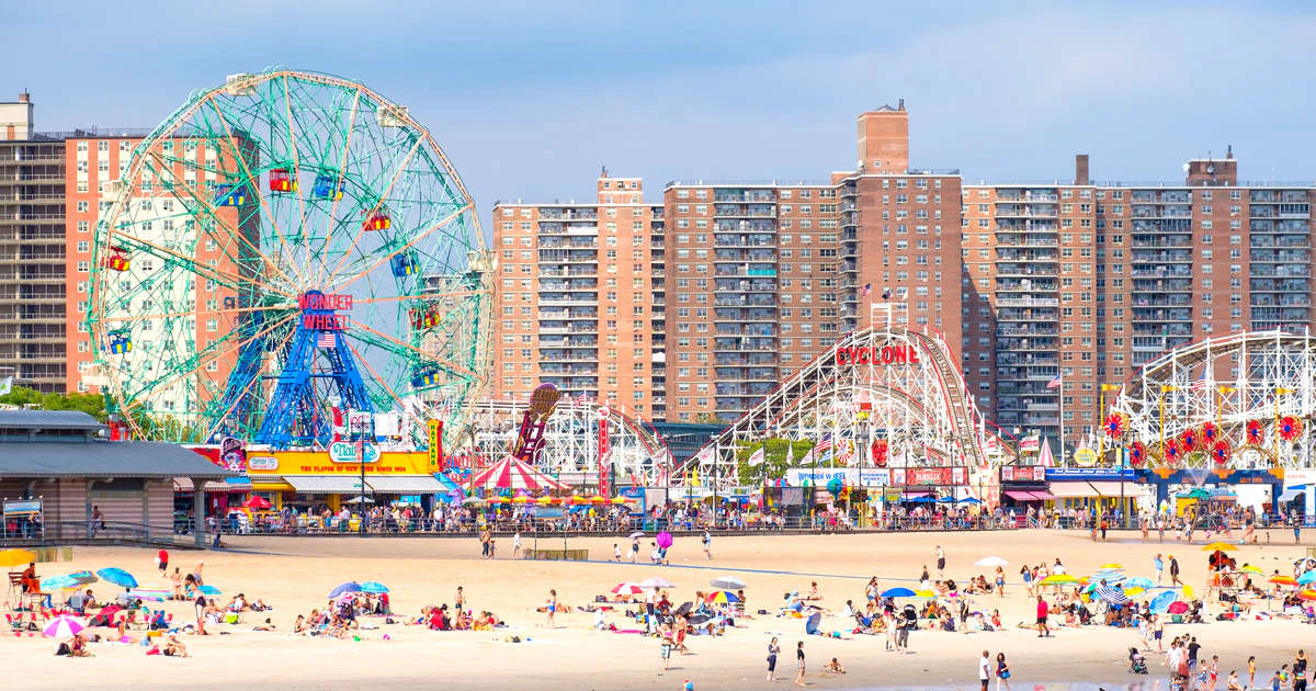 A wide view of a beach with a Ferris wheel and roller coaster in the background.