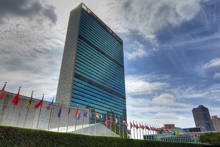 A tall building with many windows stands behind a row of flags and a hedge.