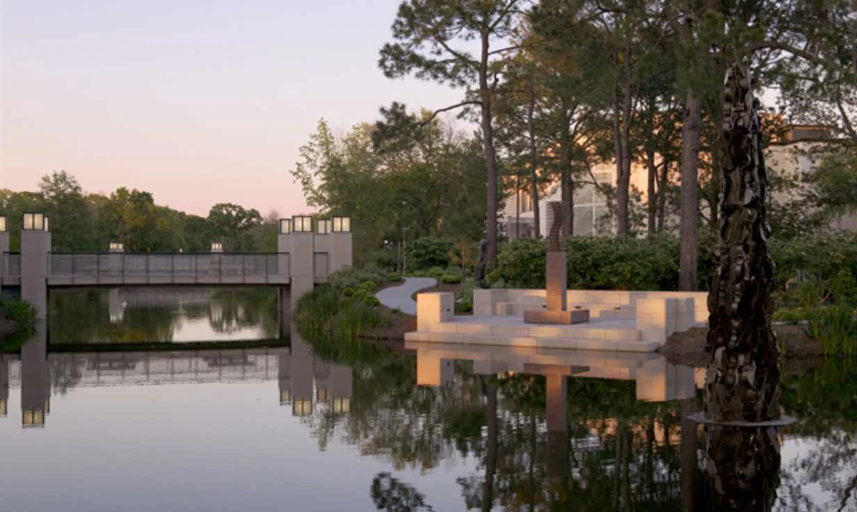 A serene outdoor space with a water feature, seating area, and bridge in the background at dusk.