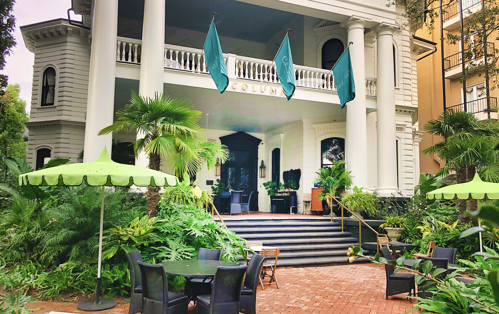 A white building with columns and a balcony, surrounded by greenery and outdoor seating.