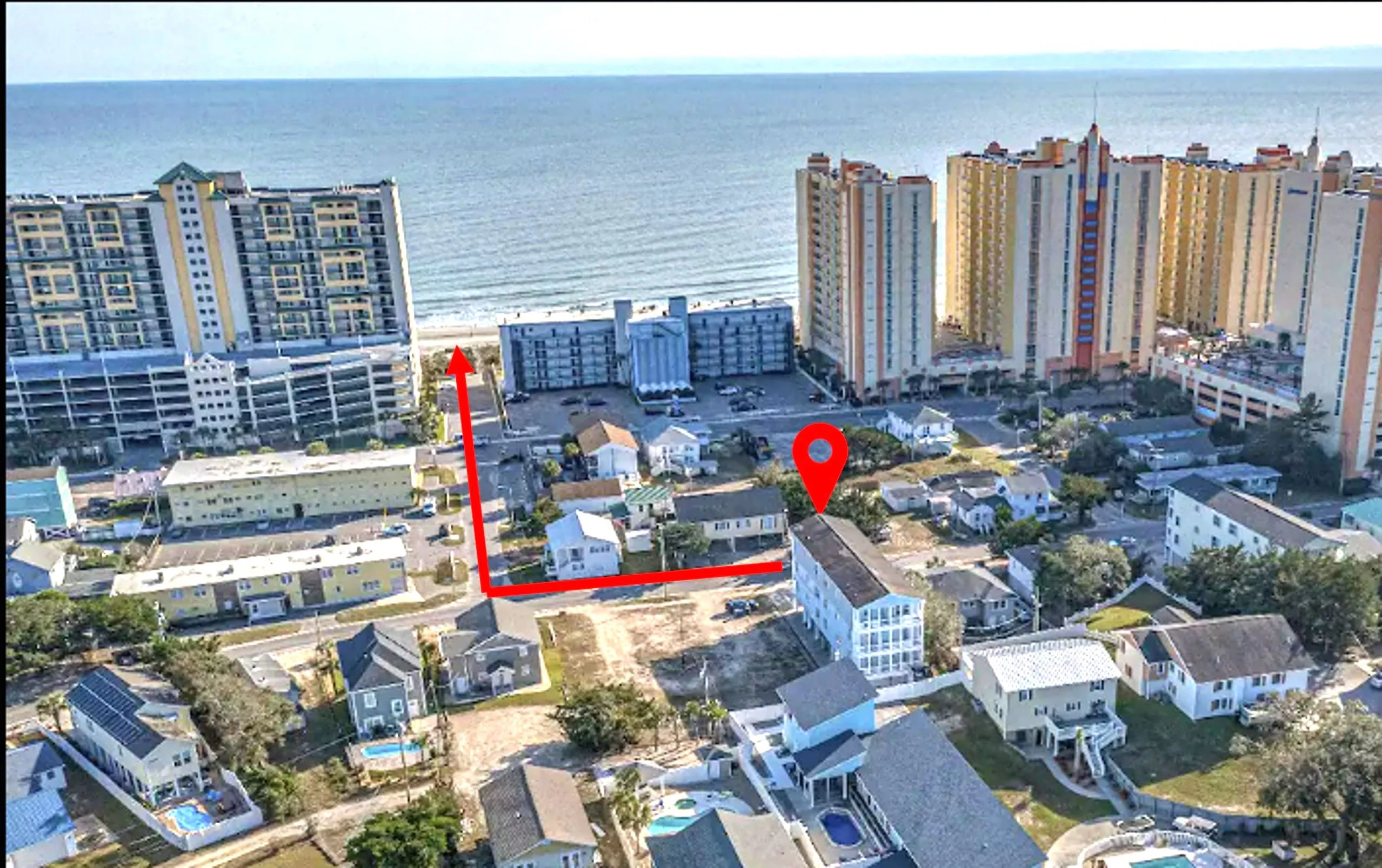 An aerial view of a beachfront property with a multi-story building, parking lot, and surrounding residential area.