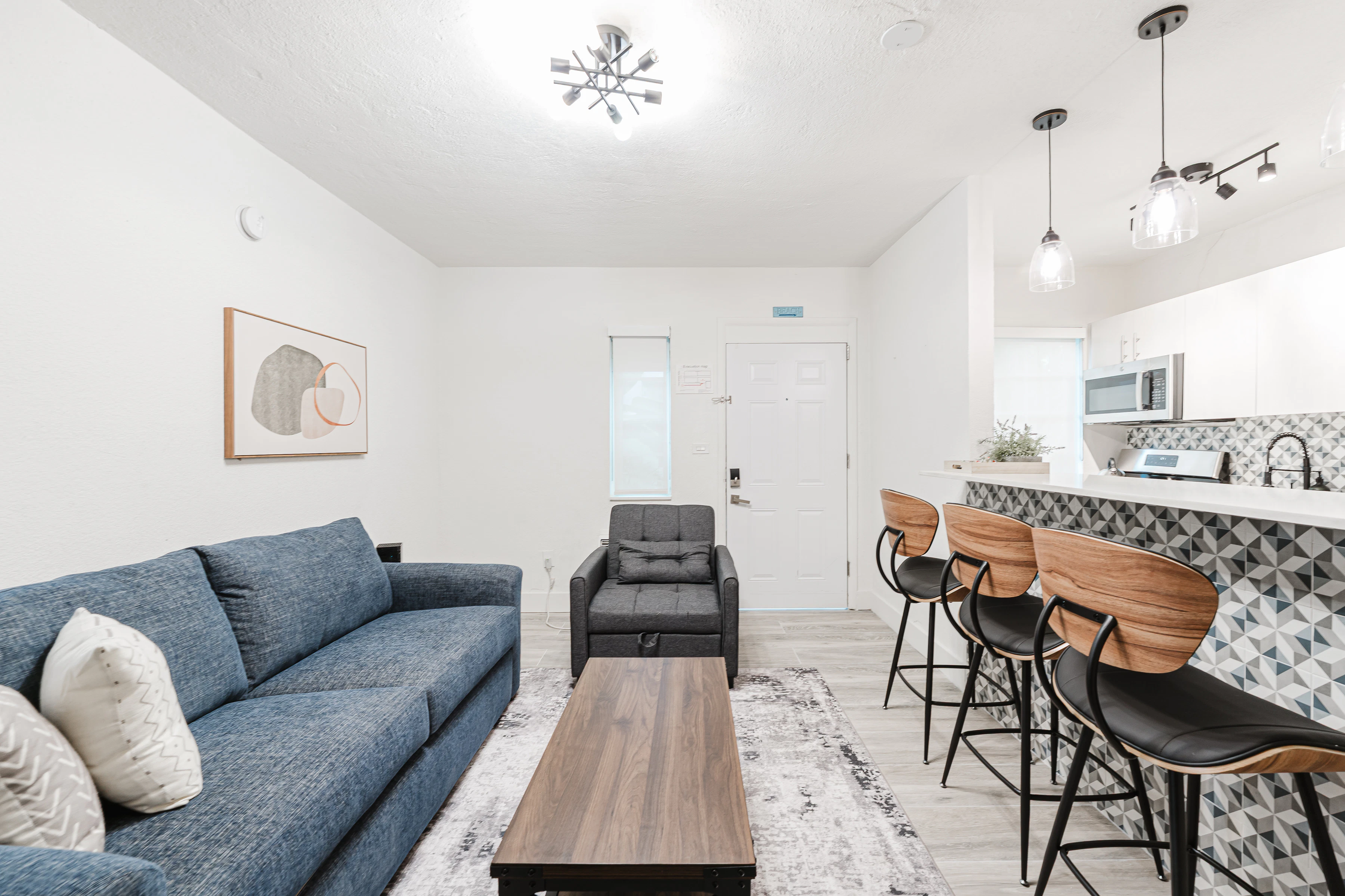 A wide view of a living room with a blue sofa, a gray armchair, a wooden coffee table, and a kitchenette with bar stools.