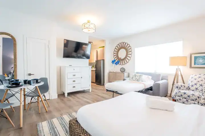 A bright and airy hotel room with a large bed, sofa, TV, dining table, and lamp.
