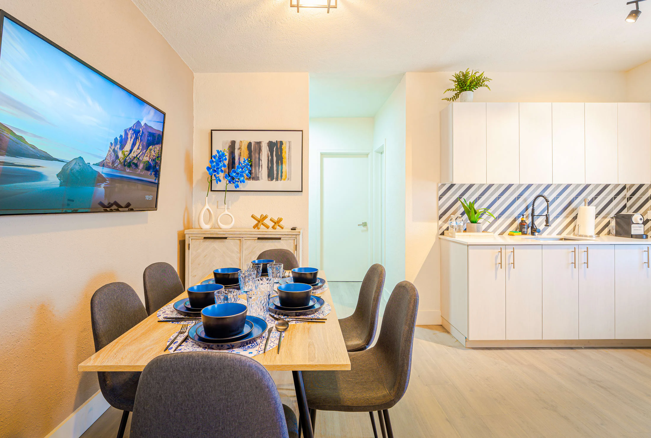 A dining area with a table set for six people, a kitchenette, a TV on the wall, and artwork.
