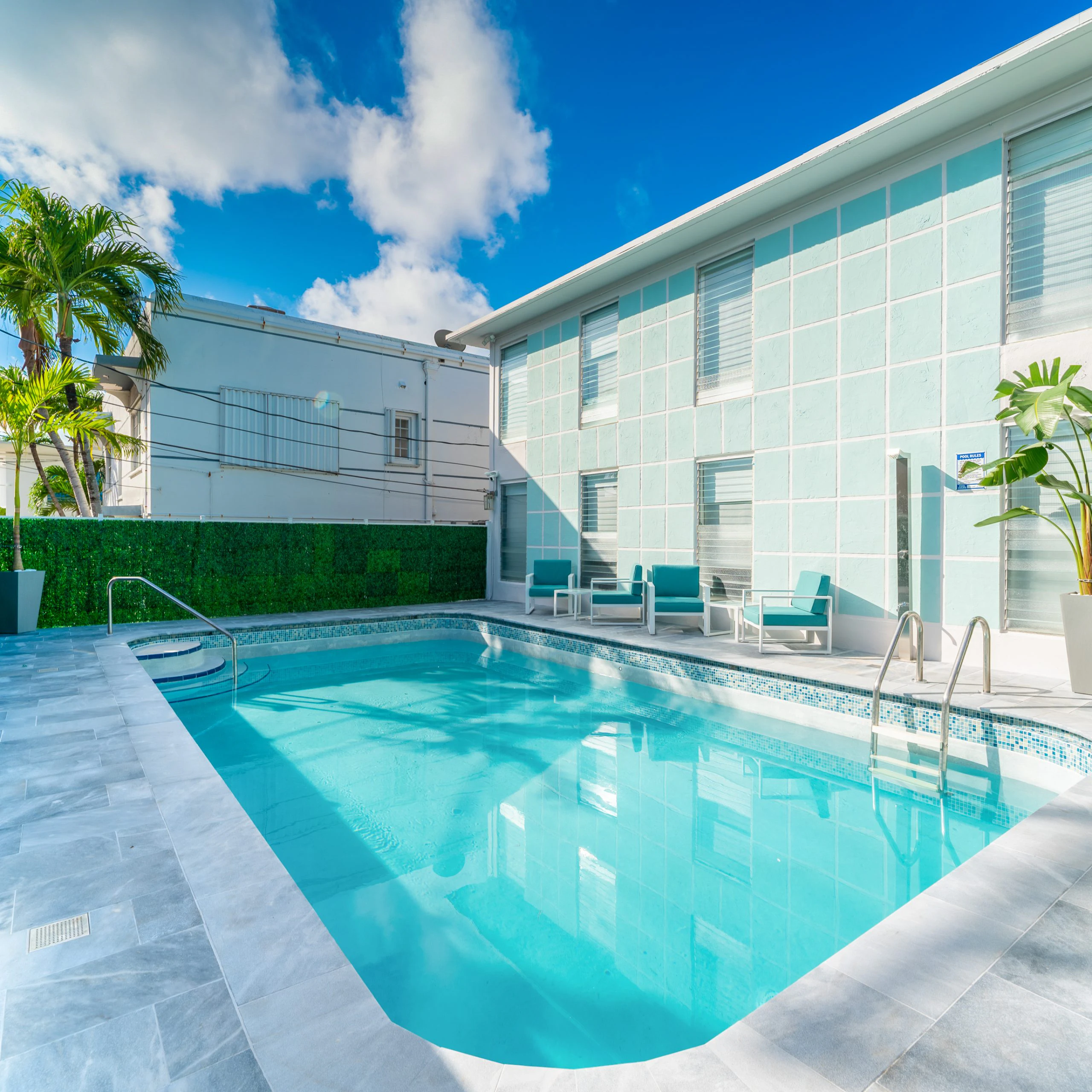 A swimming pool with a blue interior and white furniture on the side, in front of a building with large windows.