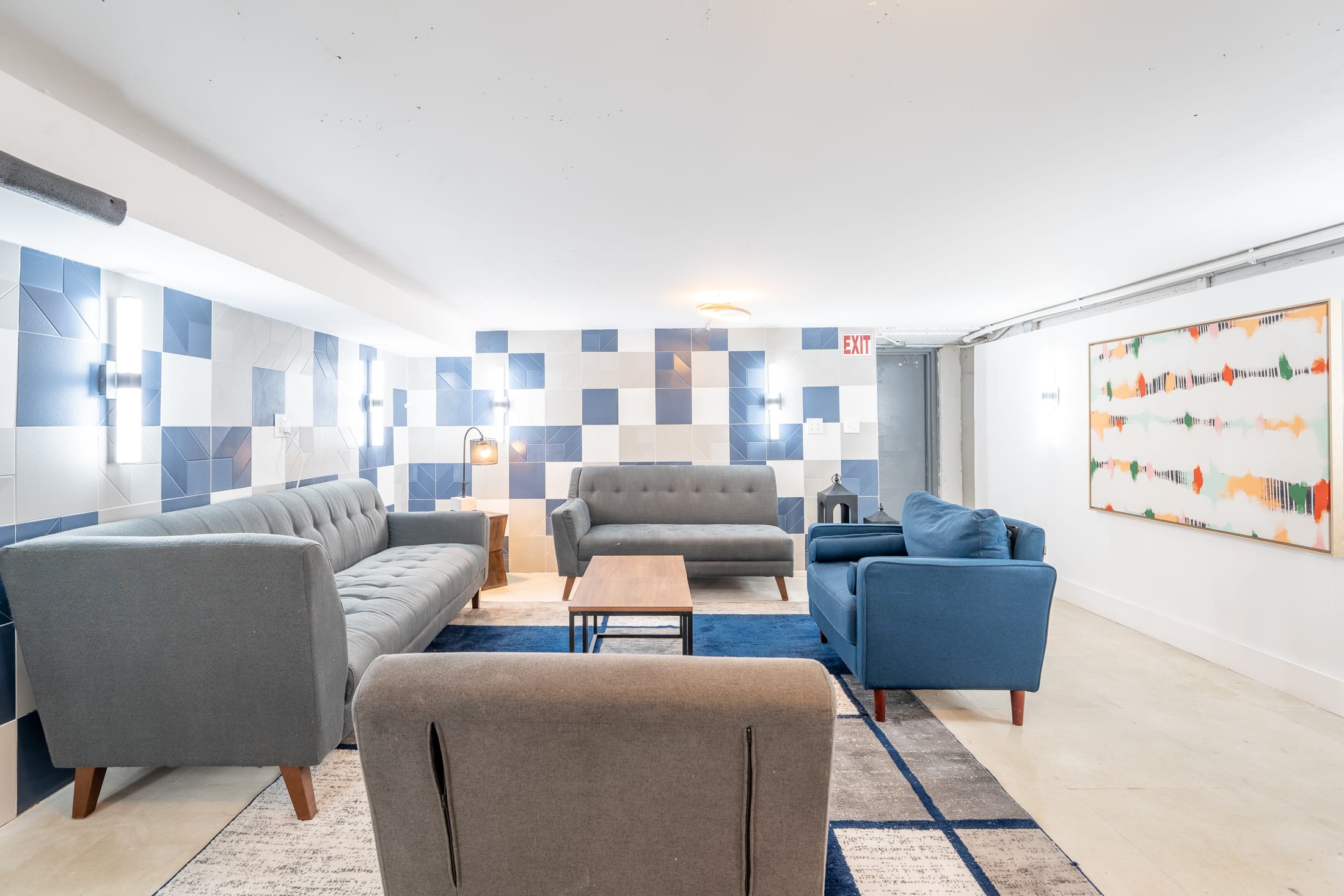 A modern lobby with gray and blue sofas and chairs, a wooden coffee table, and a colorful abstract artwork on the wall.