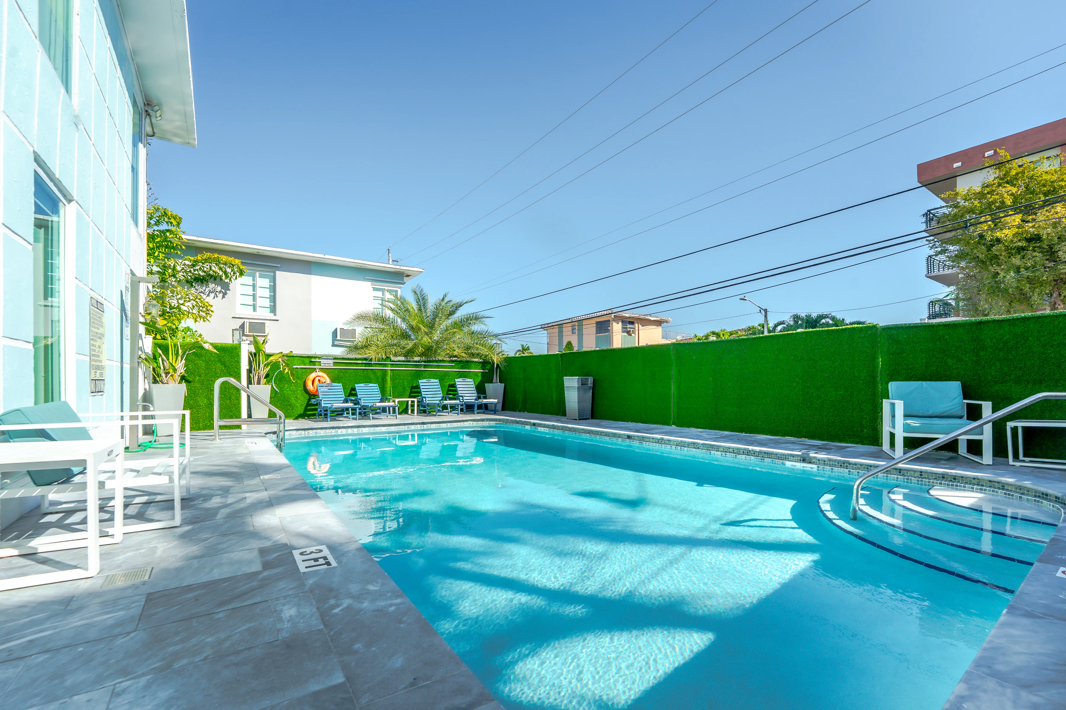 A swimming pool with lounge chairs and a green wall under a clear blue sky.