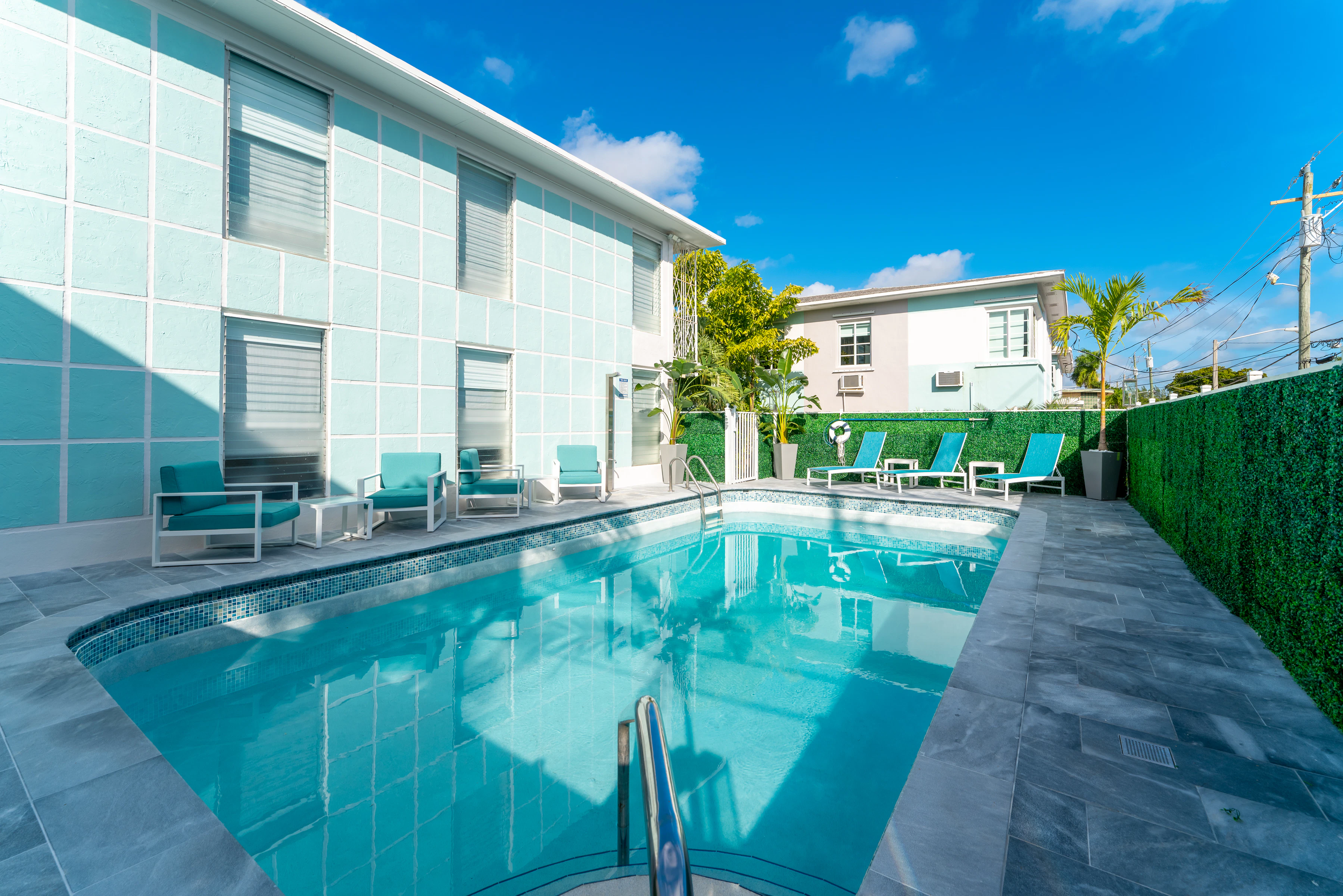 a teal pool with teal chairs and a hedge wall under a blue sky