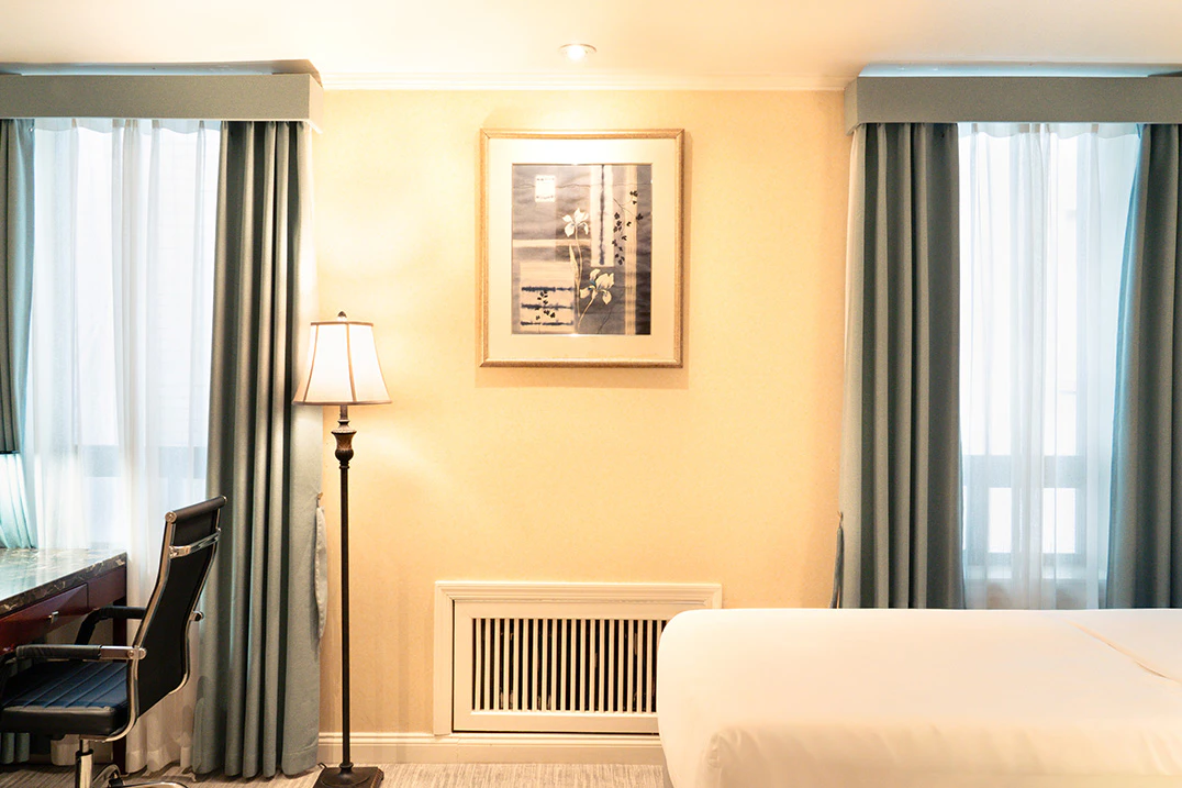 A hotel room with a bed, desk, lamp, and windows