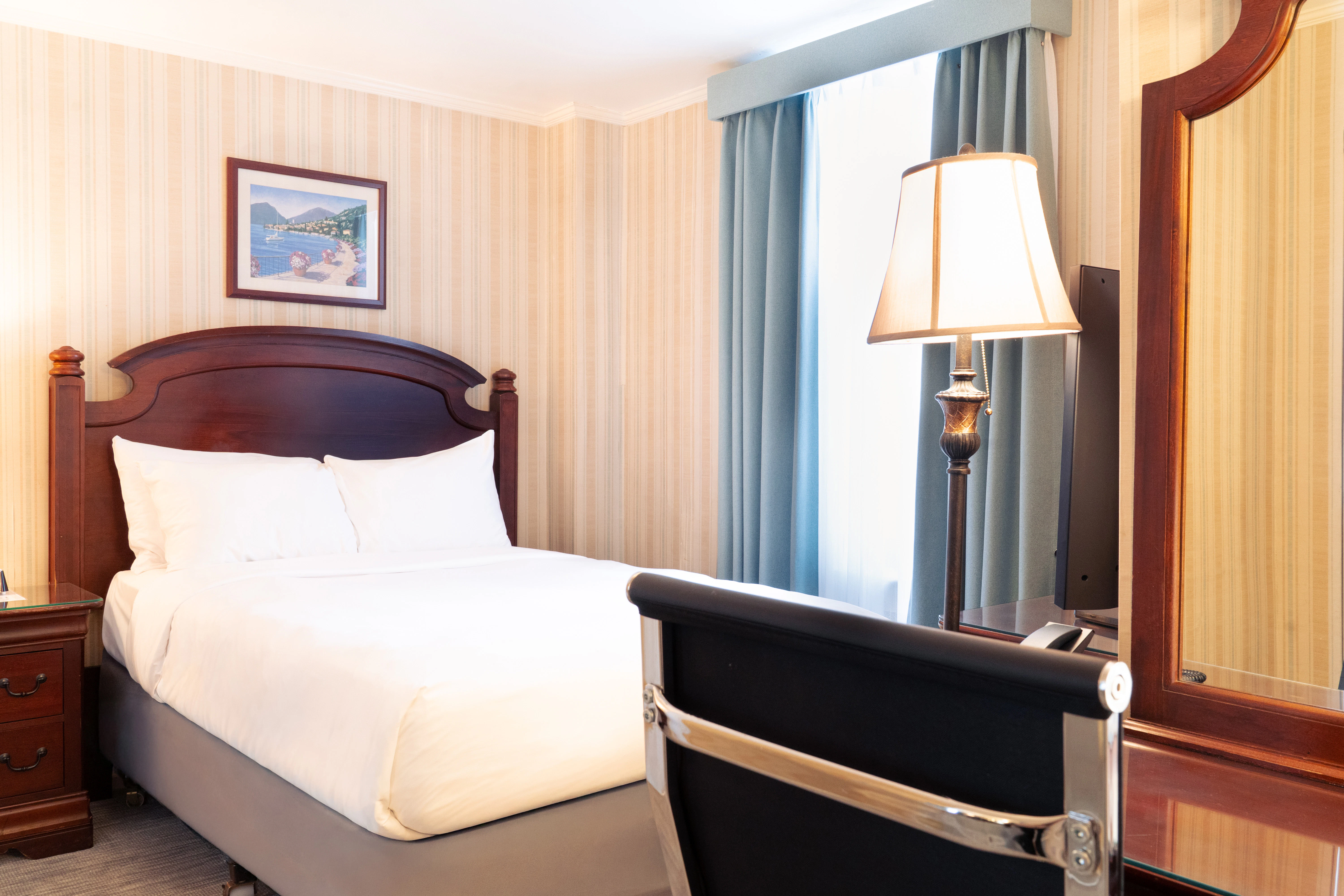 A hotel room with a bed, chair, lamp, mirror, and nightstand.