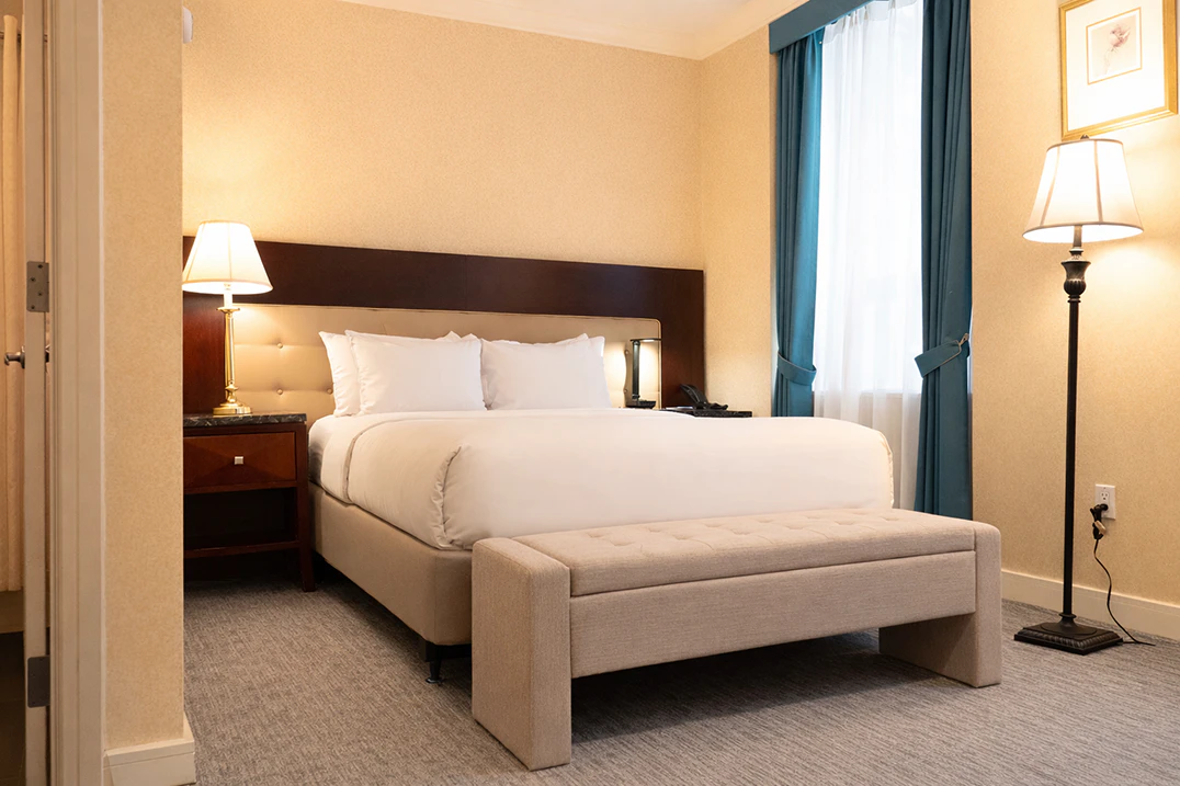 A hotel room with a large bed, beige upholstered bench, nightstand with lamp, and window with city view