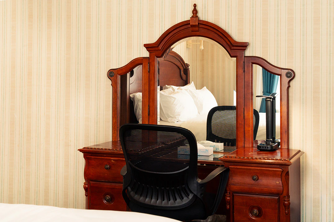 A hotel room with a large wooden dresser, mirror, and chair in front of a bed with white linens.