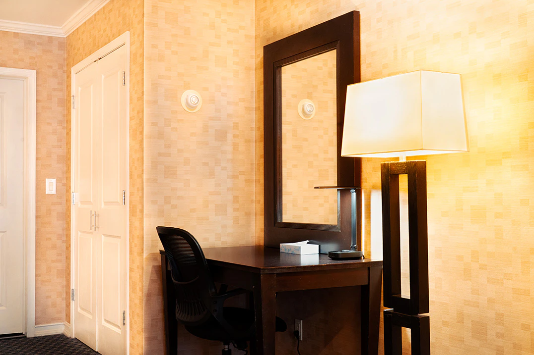 A hotel room with a desk, chair, lamp, and mirror.