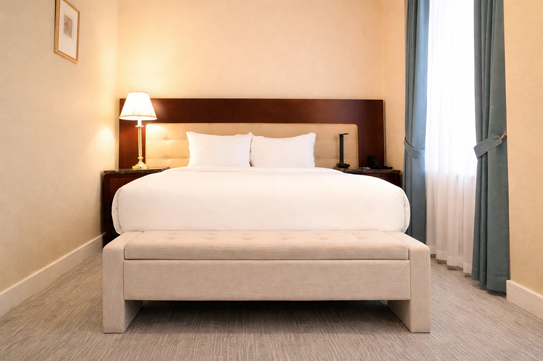 A hotel room with a large bed, beige bench, and window with curtains