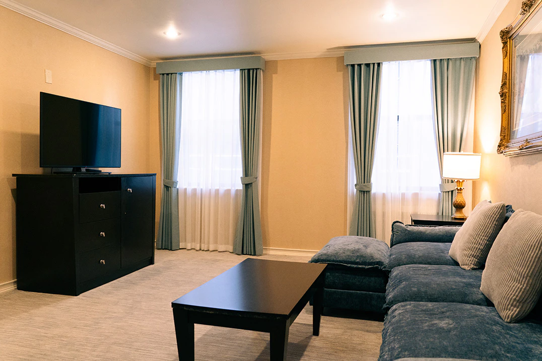 A wide view of a suite with a blue sofa, black coffee table, TV on a cabinet, and two windows with green curtains.