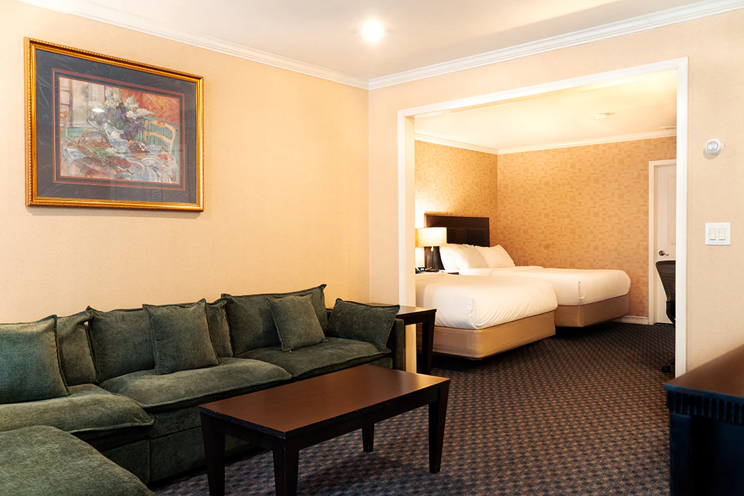 A hotel suite with a green sofa, coffee table, and a bedroom with two beds in the background.