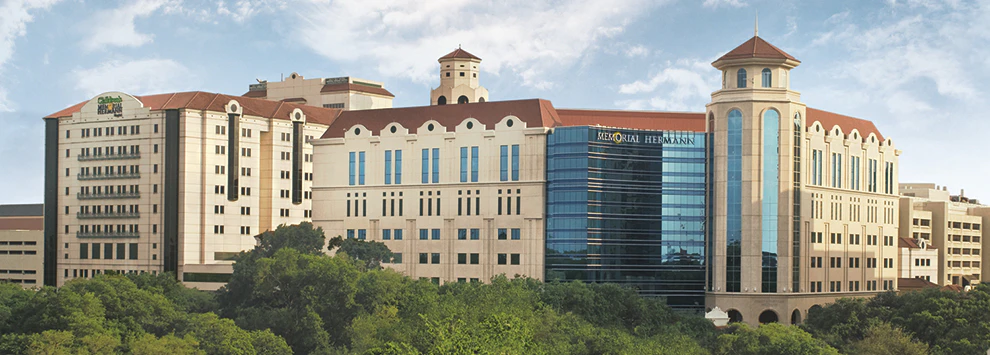A large, multi-story hotel building with a tan and brown color scheme, red-tiled roof, and glass facade, surrounded by trees under a blue sky.
