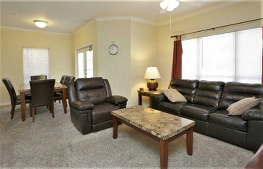 A furnished living room with a leather sofa, recliner, coffee table, dining table, and windows.