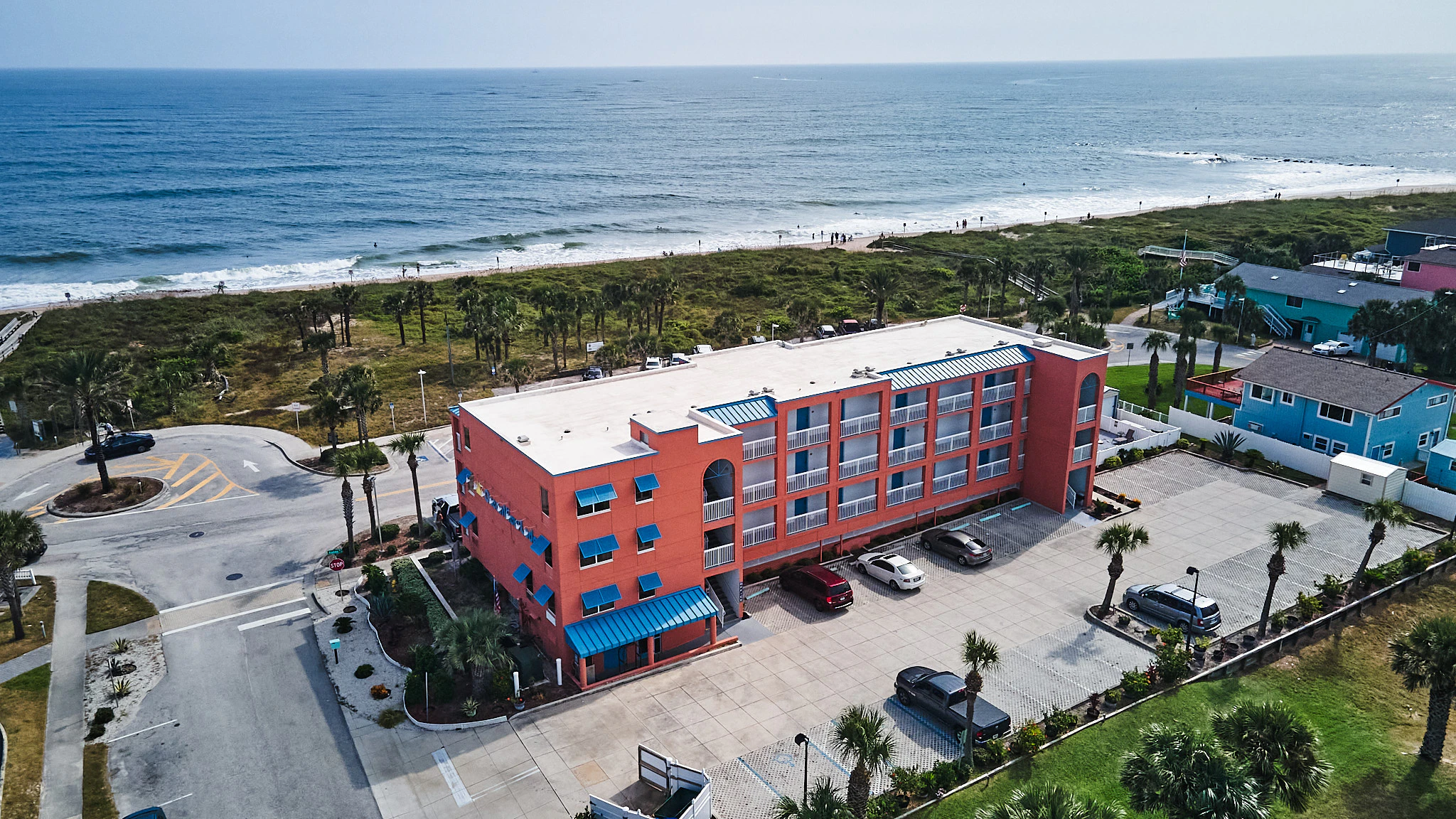 Aerial view of a red, multi-story building with blue awnings, situated near a beach with palm trees and a parking lot.